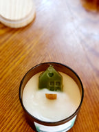 Shelter in Him scented candle featuring a decorative green house shape atop a creamy wax surface.