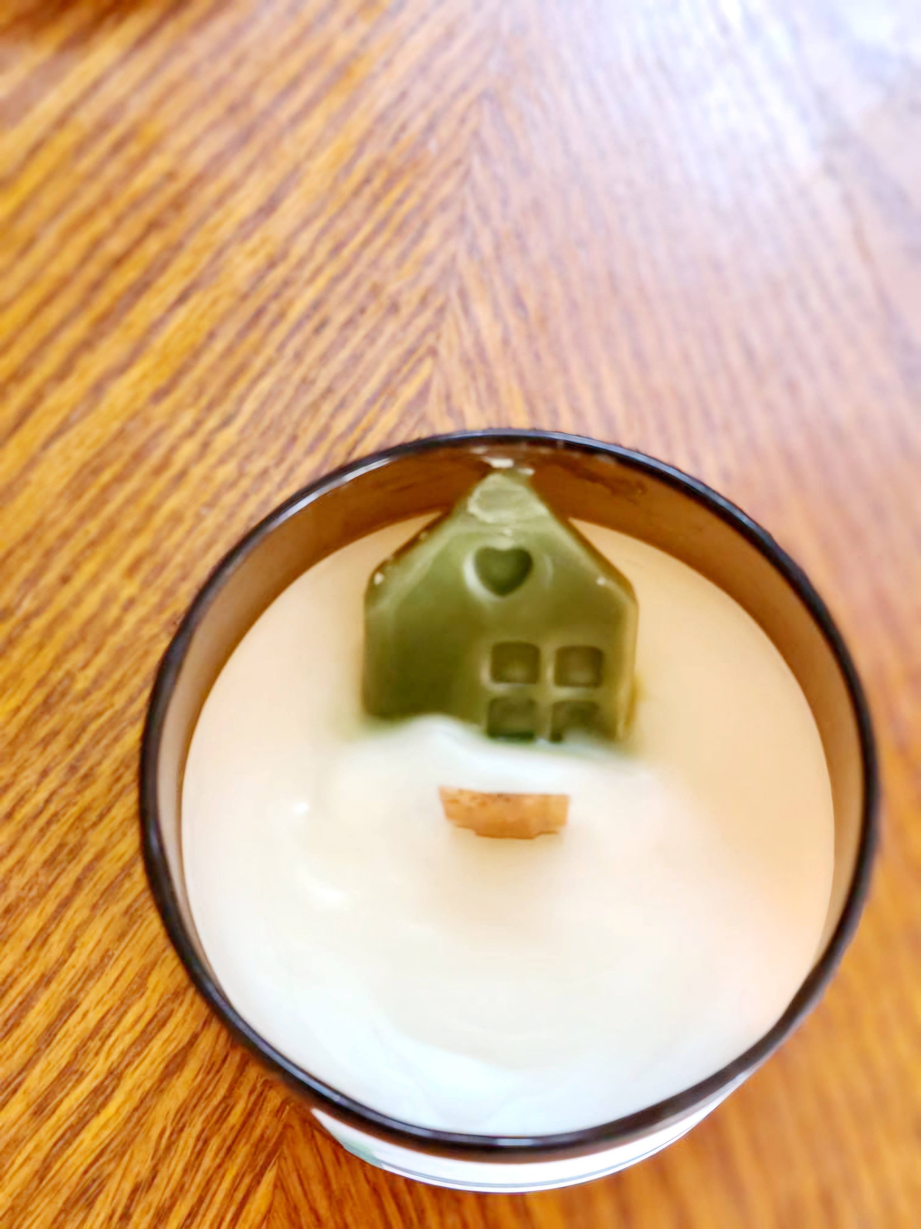 Shelter in Him candle featuring a green house shape sitting in creamy wax, evoking a campfire scent.